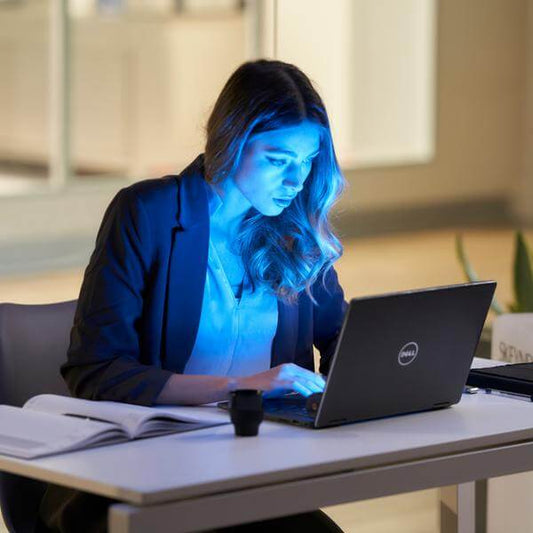 Γυναίκα εργάζεται σε laptop με έντονο μπλε φως από την οθόνη να φωτίζει το πρόσωπό της, απεικονίζοντας την έκθεση της επιδερμίδας στη μπλε ακτινοβολία κατά την πολύωρη εργασία στον υπολογιστή.