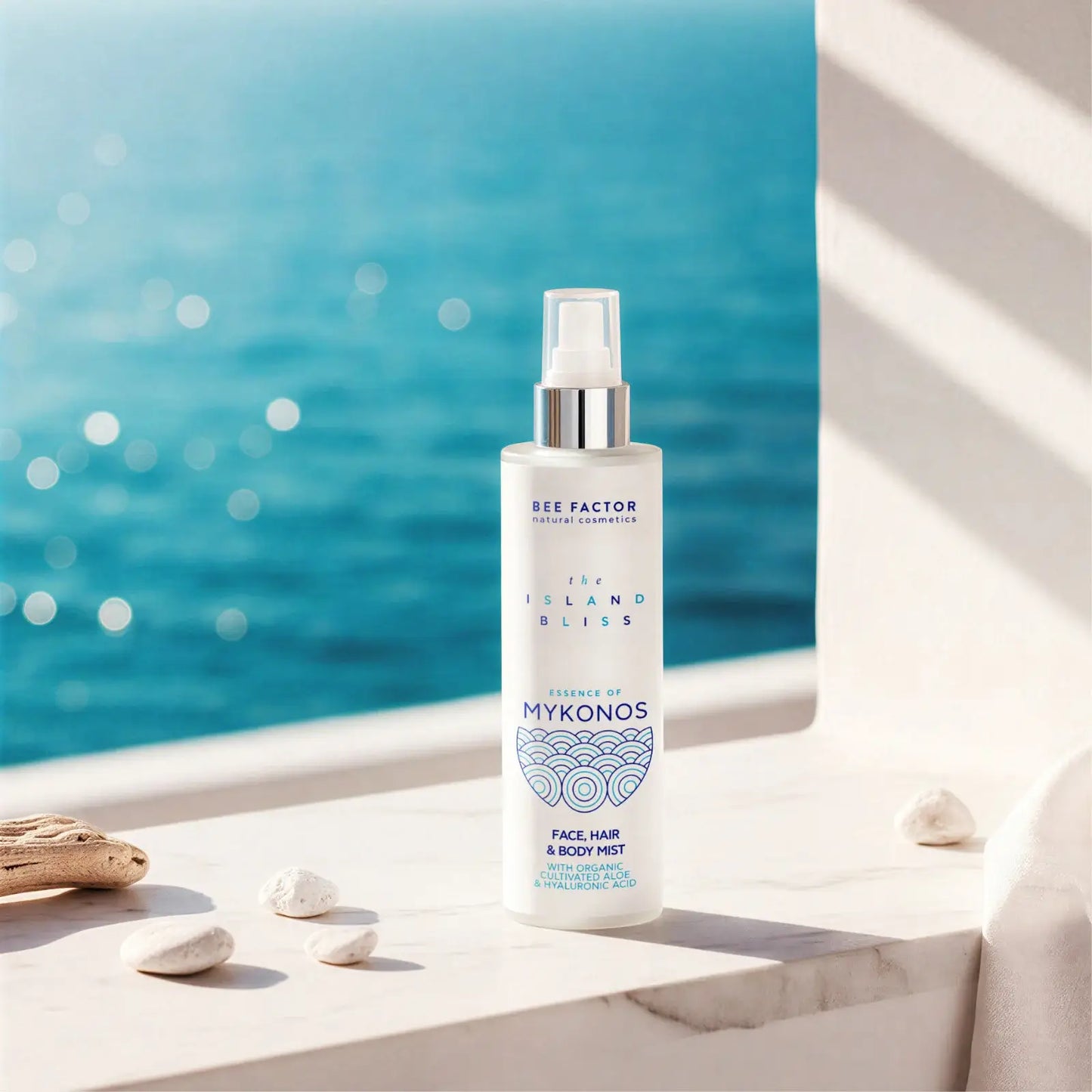 Προωθητική εικόνα του Essence of Mykonos Face, Hair & Body Mist από τη συλλογή The Island Bliss της Bee Factor Natural Cosmetics, με τη λευκή φιάλη spray να τοποθετείται σε φωτεινή μαρμάρινη επιφάνεια μπροστά από έντονο γαλάζιο θαλασσινό φόντο. Στη συσκευασία διακρίνονται το λογότυπο “Bee Factor natural cosmetics”, η ονομασία της σειράς “The Island Bliss”, η ένδειξη “Essence of Mykonos” και η κατηγορία “Face, Hair & Body Mist”, μαζί με αναφορά σε organic cultivated aloe και hyaluronic acid. Γύρω από το προϊ