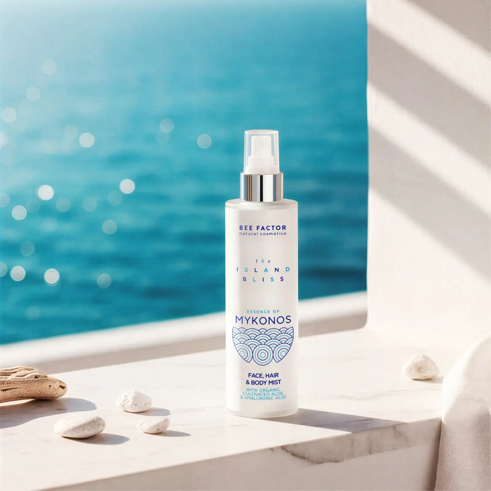 Προωθητική εικόνα του Essence of Mykonos Face, Hair & Body Mist από τη συλλογή The Island Bliss της Bee Factor Natural Cosmetics, με τη λευκή φιάλη spray να τοποθετείται σε φωτεινή μαρμάρινη επιφάνεια μπροστά από έντονο γαλάζιο θαλασσινό φόντο. Στη συσκευασία διακρίνονται το λογότυπο “Bee Factor natural cosmetics”, η ονομασία της σειράς “The Island Bliss”, η ένδειξη “Essence of Mykonos” και η κατηγορία “Face, Hair & Body Mist”, μαζί με αναφορά σε organic cultivated aloe και hyaluronic acid. Γύρω από το προϊ