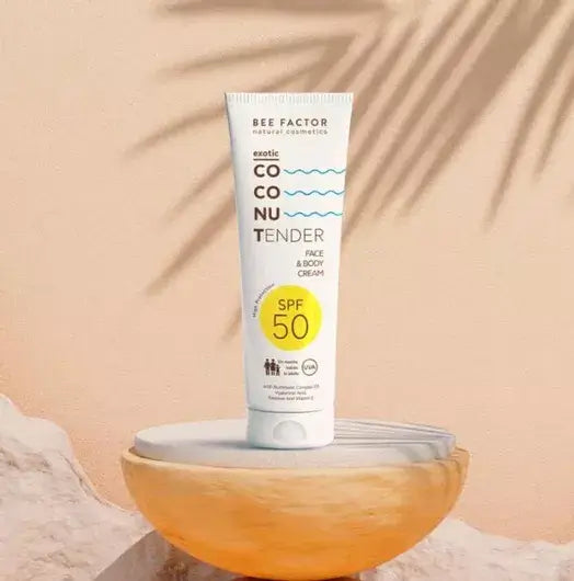 Προωθητική εικόνα της Exotic Coconut Tender Cream SPF 50 της Bee Factor Natural Cosmetics, με τη λευκή σωληναριακή συσκευασία να στέκεται όρθια πάνω σε γυάλινο διακοσμητικό μπολ σε ζεστό μπεζ φόντο. Στη συσκευασία διακρίνονται το λογότυπο “Bee Factor natural cosmetics”, η ονομασία “Exotic Coconut Tender”, η ένδειξη “Face & Body Cream” και το έντονο σήμα “SPF 50”, ενώ η σύνθεση της εικόνας συμπληρώνεται από σκιές φύλλων φοίνικα και φυσικά βραχώδη στοιχεία που παραπέμπουν σε καλοκαιρινή, τροπική αισθητική. Η 