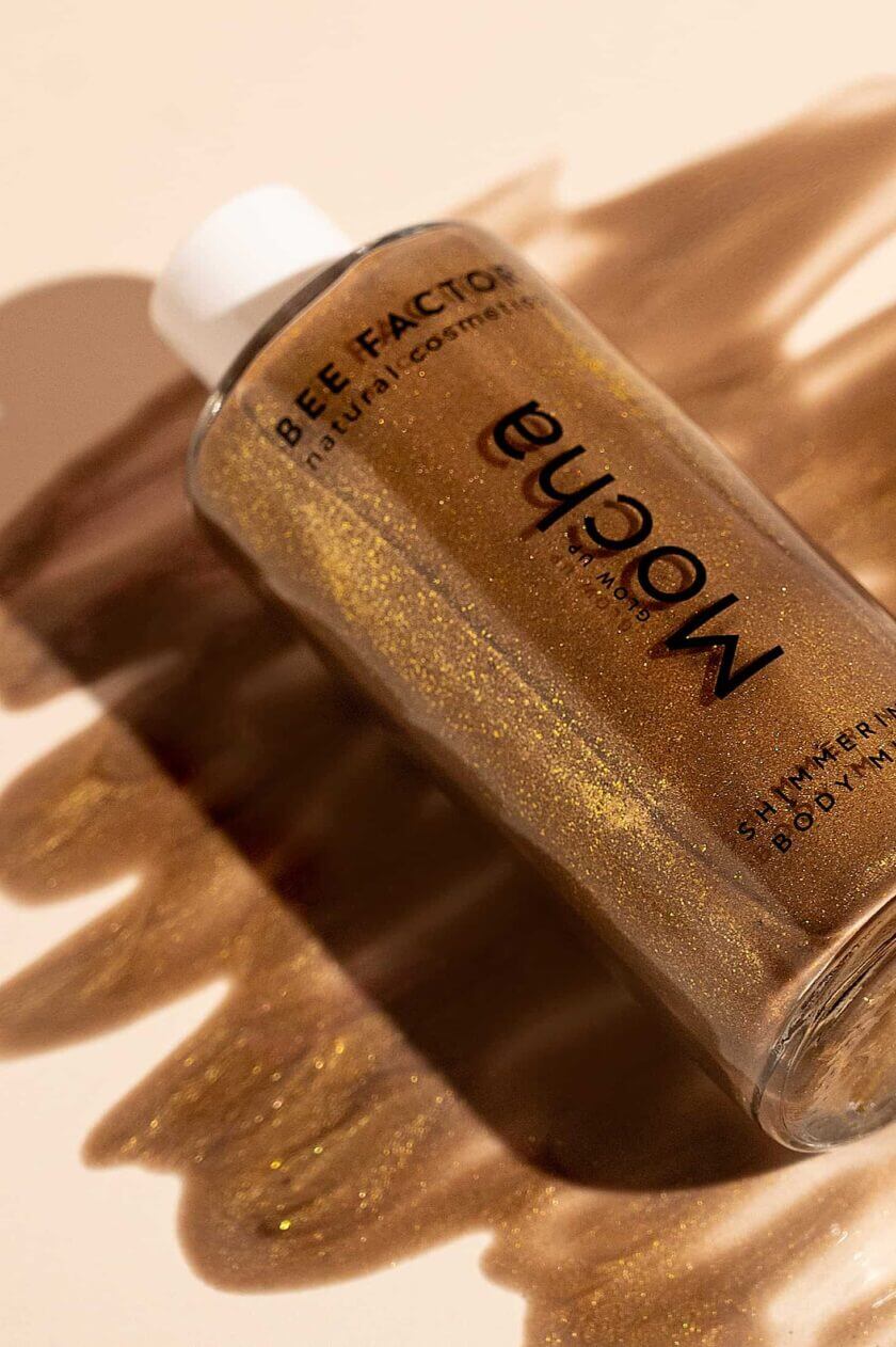 Μπουκάλι Glow Up Mocha Shimmering Body Milk με φώτο το προϊόν απλωμένο της Bee Factor. Ενυδατικό γαλάκτωμα σώματος με χρυσές χρωστικές που προσφέρει λαμπερή bronze λάμψη και αναδεικνύει την επιδερμίδα. Θρέφει το δέρμα με aloe vera, φυτική γλυκερίνη και πανθενόλη, ενώ αφήνει το δέρμα απαλό και μεταξένιο, χωρίς να αφήνει λιπαρότητα ή λεκέδες. Ένα must-have για εφέ μαυρίσματος και μοναδικό άρωμα ανθών βανίλιας, περγαμόντο και μανόλιας. Διαθέσιμο στο Eiria Cosmetics.