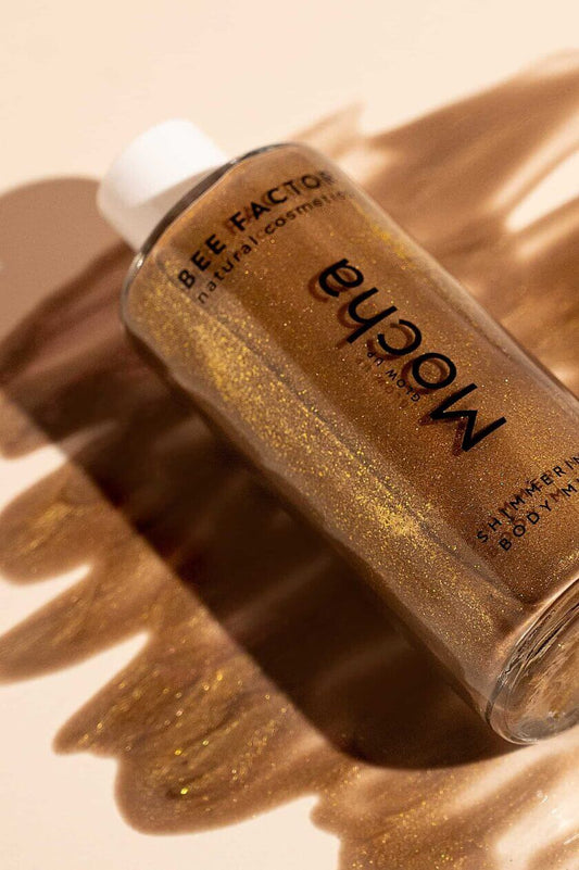 Μπουκάλι Glow Up Mocha Shimmering Body Milk με φώτο το προϊόν απλωμένο της Bee Factor. Ενυδατικό γαλάκτωμα σώματος με χρυσές χρωστικές που προσφέρει λαμπερή bronze λάμψη και αναδεικνύει την επιδερμίδα. Θρέφει το δέρμα με aloe vera, φυτική γλυκερίνη και πανθενόλη, ενώ αφήνει το δέρμα απαλό και μεταξένιο, χωρίς να αφήνει λιπαρότητα ή λεκέδες. Ένα must-have για εφέ μαυρίσματος και μοναδικό άρωμα ανθών βανίλιας, περγαμόντο και μανόλιας. Διαθέσιμο στο Eiria Cosmetics.