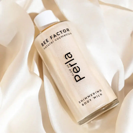 Το Glow Up Perla Shimmering Body Milk της Bee Factor Natural Cosmetics σε διάφανη φιάλη με λευκό καπάκι, τοποθετημένο λοξά πάνω σε απαλό σατινέ ύφασμα σε εκρού και λευκές αποχρώσεις. Η συσκευασία αναδεικνύει την περλέ, λαμπερή σύνθεση του προϊόντος, ενώ στην ετικέτα διακρίνονται το λογότυπο “Bee Factor natural cosmetics”, η ονομασία “Perla” και η ένδειξη “Shimmering Body Milk”. Η εικόνα αποπνέει αίσθηση κομψότητας, θηλυκότητας και πολυτελούς περιποίησης σώματος.