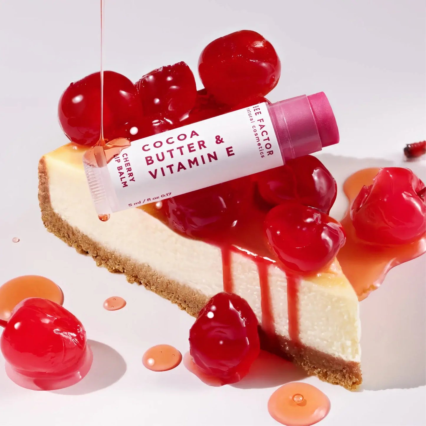 Bee Factor cherry lip balm (ενυδατικό βάλσαμο χειλιών με γεύση κεράσι) 5ml με cocoa butter & vitamin E, τοποθετημένο πάνω σε κομμάτι cheesecake με γυαλιστερό σιρόπι και κεράσια σε λευκό φόντο – Eiria.gr