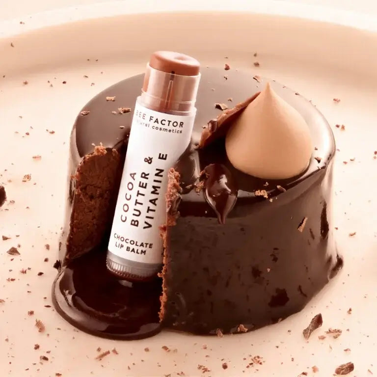 Δημιουργική προωθητική εικόνα του Bee Factor Chocolate Lip Balm, με το lip balm σε λευκή stick συσκευασία τοποθετημένο μέσα σε σοκολατένιο γλυκό με ρευστή σοκολάτα. Το προϊόν αναγράφει “Cocoa Butter & Vitamin E” και ξεχωρίζει στο κέντρο της σύνθεσης, περιτριγυρισμένο από λαχταριστή σοκολατένια υφή, κομμάτια κέικ και κρέμα σε ανοιχτή καραμελέ απόχρωση στην κορυφή. Η εικόνα συνδέει οπτικά το προϊόν με το άρωμα σοκολάτας και αποπνέει αίσθηση πλούσιας περιποίησης και απόλαυσης.