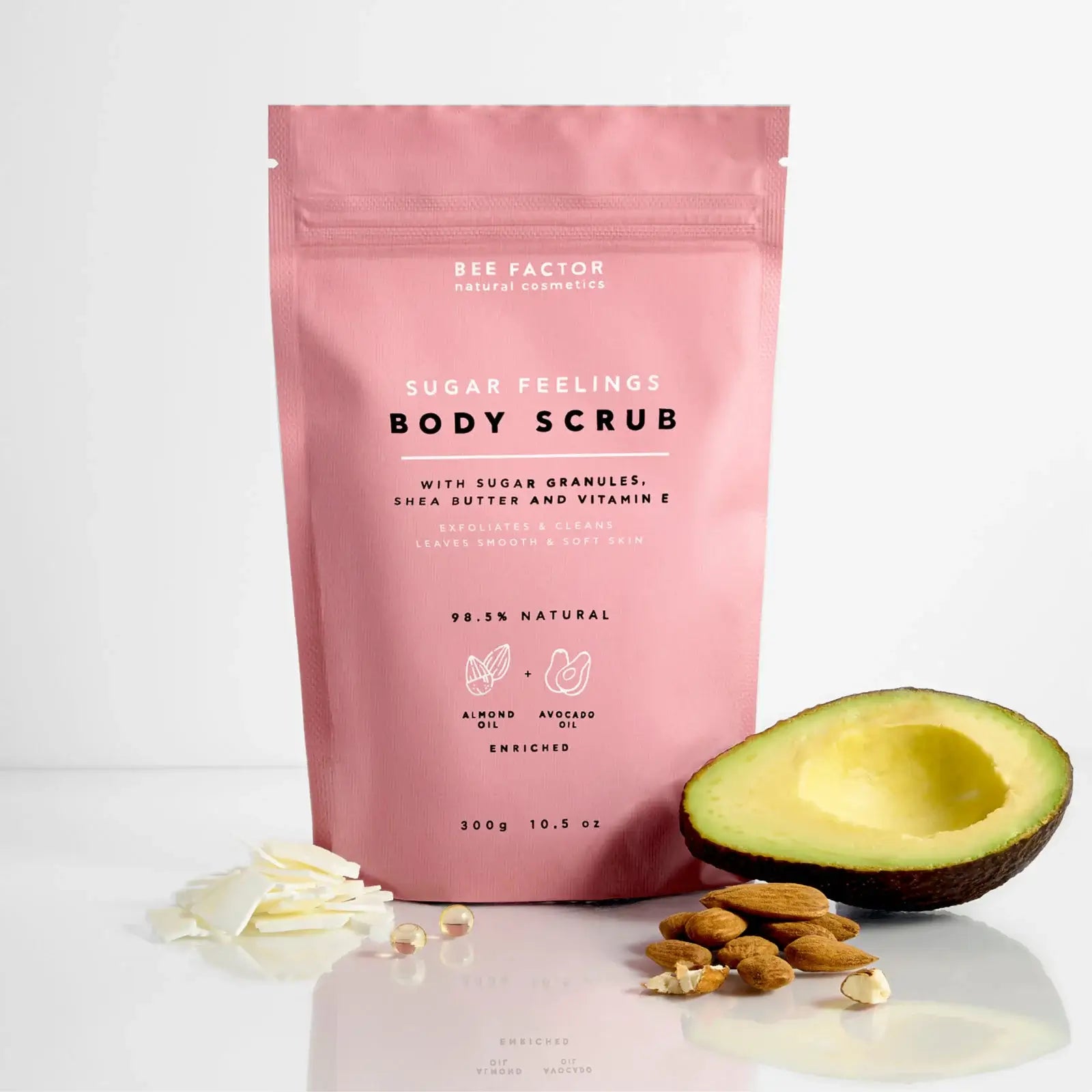 Το Sugar Feelings Body Scrub της Bee Factor Natural Cosmetics σε ροζ συσκευασία τύπου pouch με επανακλειόμενο κλείσιμο, τοποθετημένο σε καθαρό ανοιχτόχρωμο φόντο. Μπροστά από τη συσκευασία εμφανίζονται μισό αβοκάντο, αμύγδαλα, λευκά flakes βουτύρου καριτέ και κάψουλες βιταμίνης Ε, στοιχεία που αναδεικνύουν τα θρεπτικά συστατικά της σύνθεσης. Στην μπροστινή όψη αναγράφονται η ονομασία “Sugar Feelings Body Scrub”, η σύνθεση με κόκκους ζάχαρης, βούτυρο καριτέ και βιταμίνη Ε, καθώς και η ένδειξη ότι το προϊόν ε