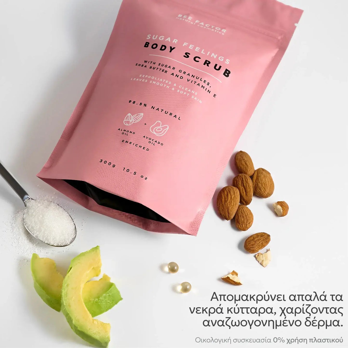 Προωθητική εικόνα του Sugar Feelings Body Scrub της Bee Factor, με τη ροζ συσκευασία του προϊόντος τοποθετημένη λοξά σε λευκό φόντο, πλαισιωμένη από ζάχαρη σε κουτάλι, φέτες αβοκάντο, αμύγδαλα και κάψουλες ελαίου που παραπέμπουν στα φυσικά συστατικά της σύνθεσης. Στη συσκευασία αναγράφονται η ονομασία του προϊόντος, η σύνθεση με κόκκους ζάχαρης, βούτυρο καριτέ και βιταμίνη Ε, καθώς και η ένδειξη ότι είναι 98,5% φυσικό και εμπλουτισμένο με αμυγδαλέλαιο και έλαιο αβοκάντο. Στο κάτω μέρος της εικόνας τονίζεται